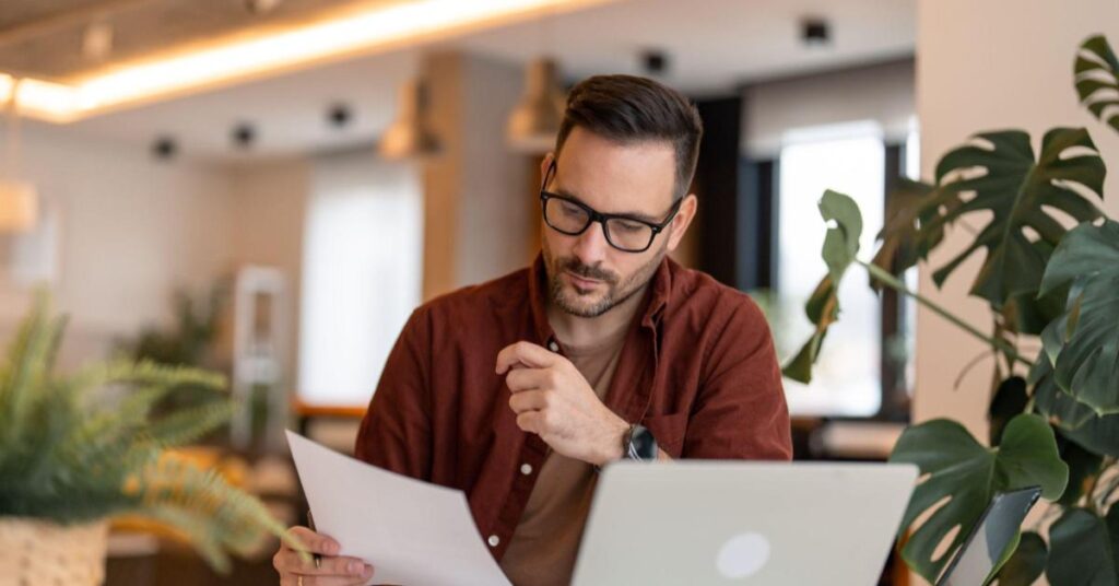 A imagem mostra uma sala iluminada com diversas plantas, e um homem sentado à mesa, escrevendo. Ele é branco, tem barba e cabelos curtos, usa óculos preto quadrado e veste uma camisa vinho. A imagem representa o momento de planejamento na estratégia de marketing de conteúdo.
